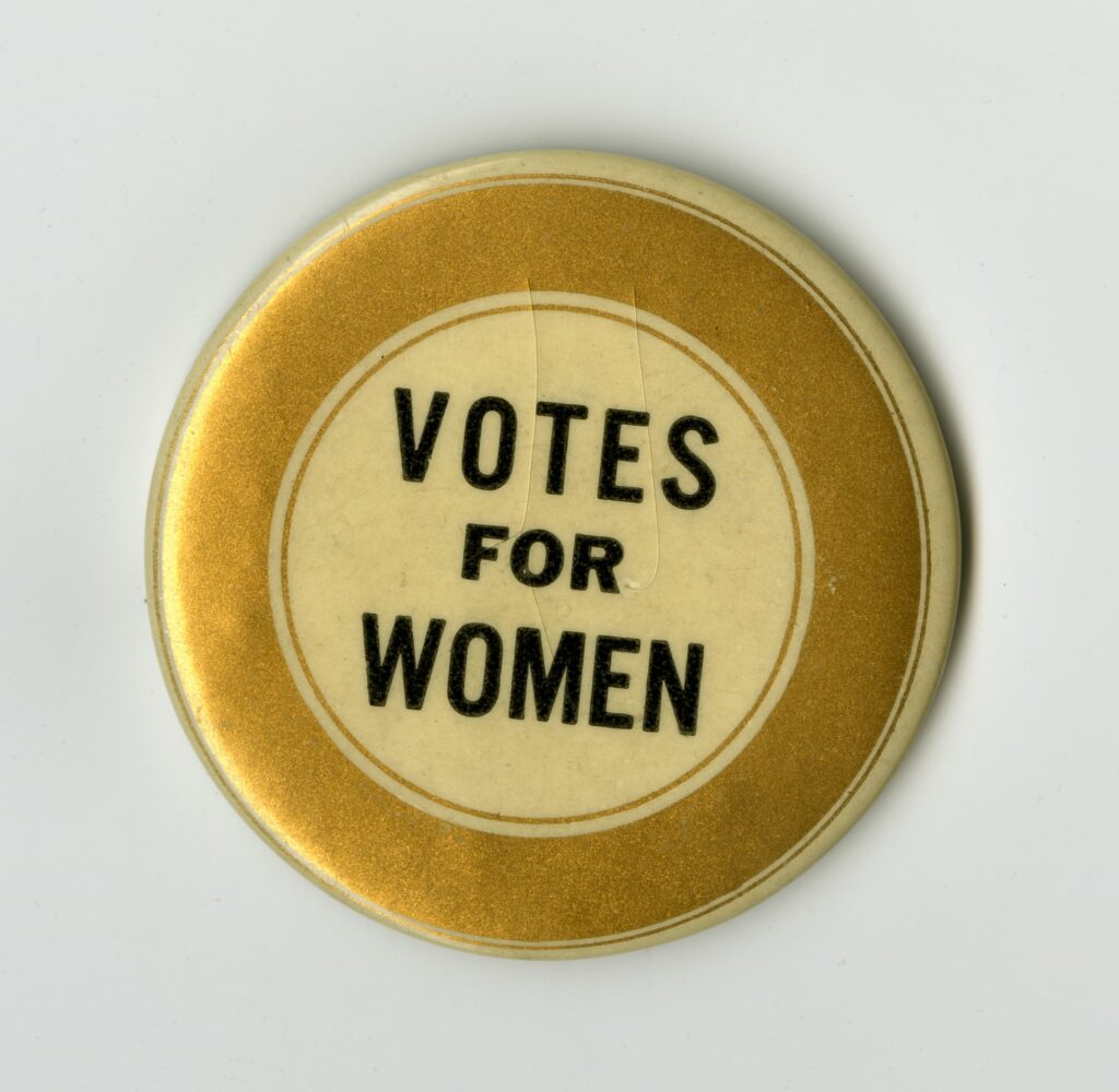 Color photo of a button from the early 1900s supporting women's suffrage that says "Votes for Women" in black text surrounded by a gold circle border; Public domain image, courtesy of RawPixel.com